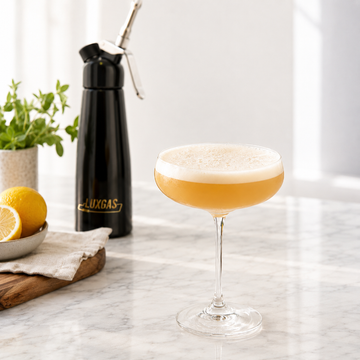 Cocktail with a fine foam layer beside a professional cream siphon on a bar counter